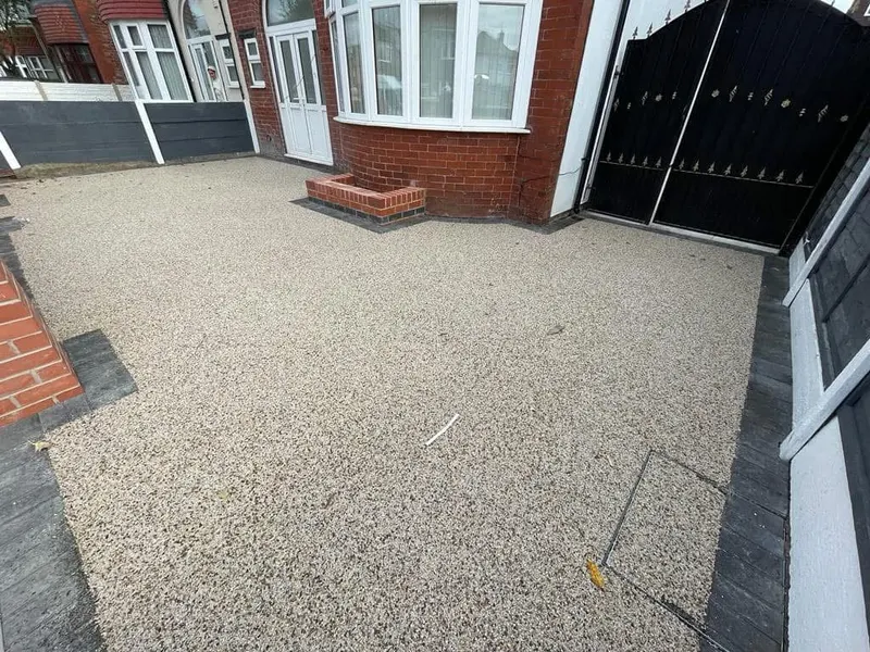 Full-width light cream resin bound front area with charcoal block borders, a built-in brick planter, and a resin-filled manhole beside ornate metal gates.