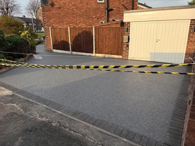 Pale grey resin bound driveway with charcoal block borders in front of a white garage door, blocked off with yellow and black warning tape.