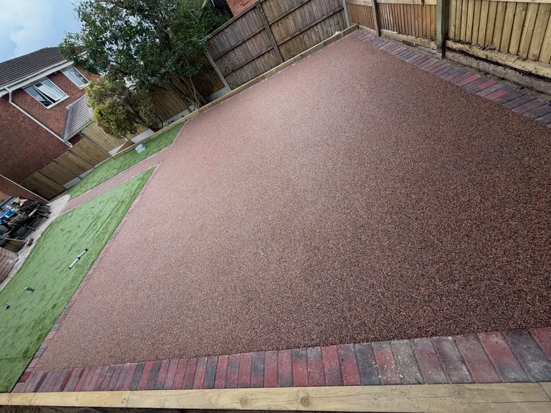 Warm reddish-brown resin bound patio and narrow path with multi-colour block borders, artificial grass, timber edging, and panel fencing.