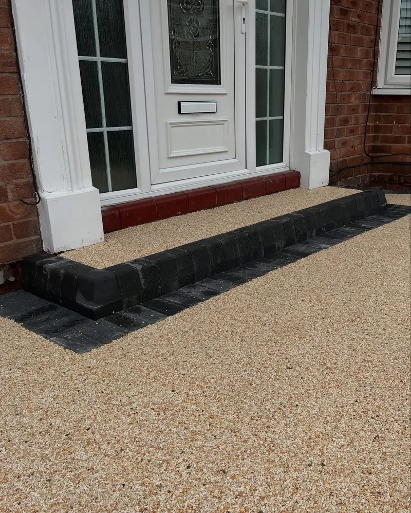 Golden-buff resin bound entrance area with an L-shaped charcoal block step and border in front of a white door on red brick walls.