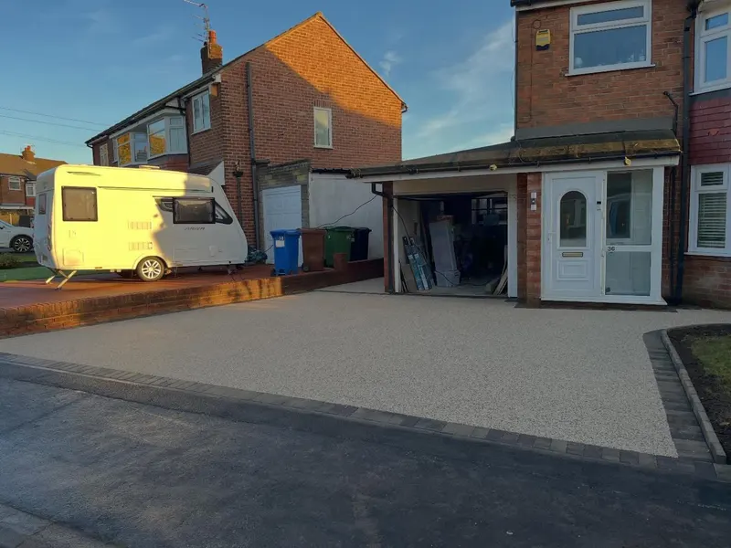 Light cream resin bound driveway with charcoal block borders across a red brick semi-detached front, beside an open garage and a parked caravan.