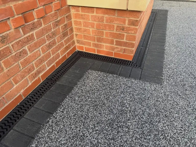Silver-grey resin bound surfacing meeting a charcoal block border and black channel drain along the base of a red brick house wall.