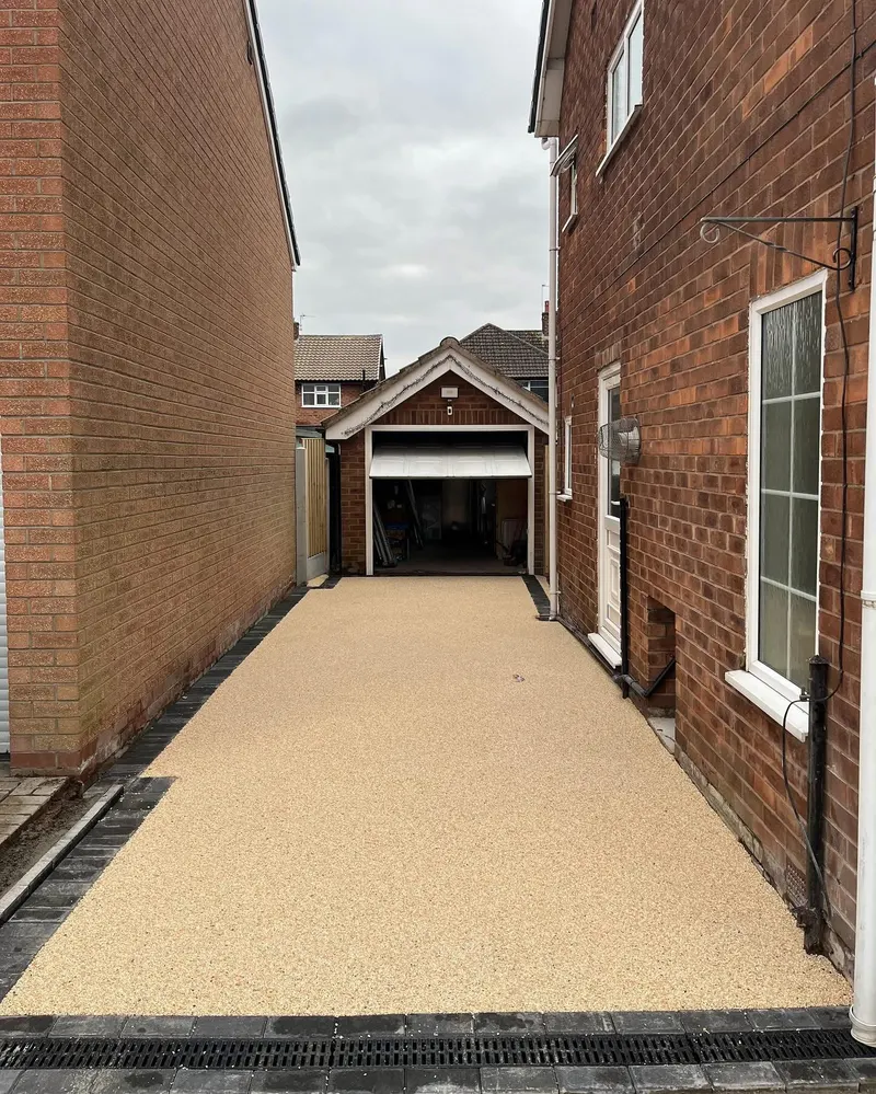 Straight sandy-beige resin bound driveway with dark block borders on both sides, a front drainage grate, leading to a small brick garage.