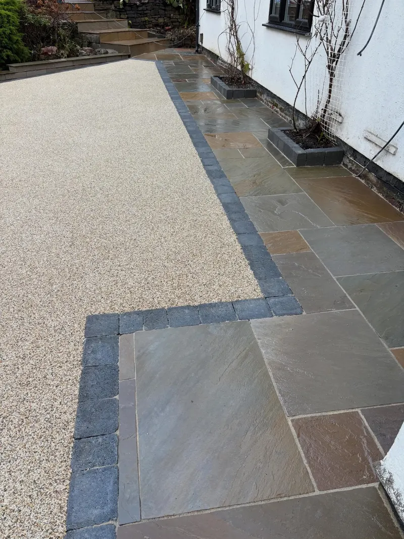 Light beige resin bound area separated from multi-tonal natural stone paving by a charcoal block border, with block planters against a white wall.