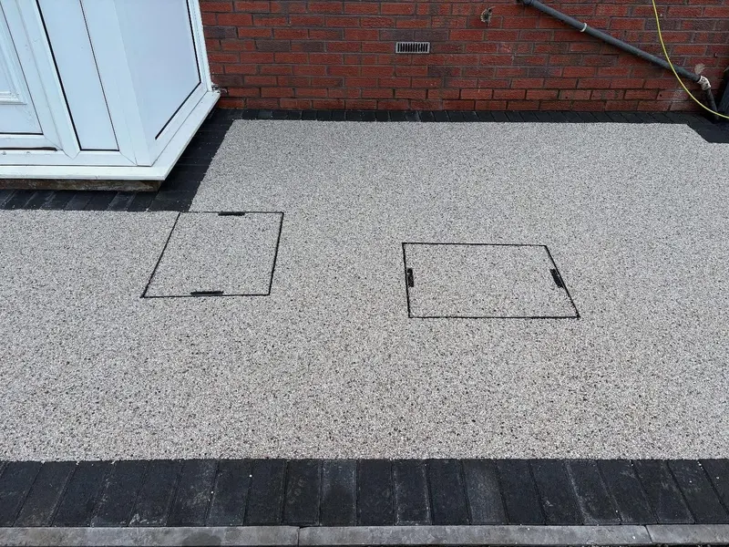 Light grey resin bound surface beside a red brick wall, with two recessed covers infilled to match and a charcoal block paving border.
