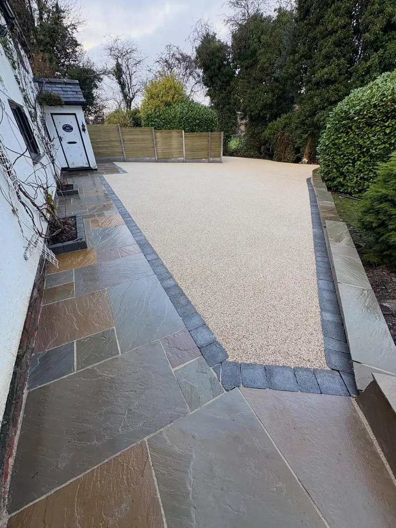 Large cream resin bound section bordered by dark setts and surrounded by mixed-colour natural stone paving beside a white house and garden fence.