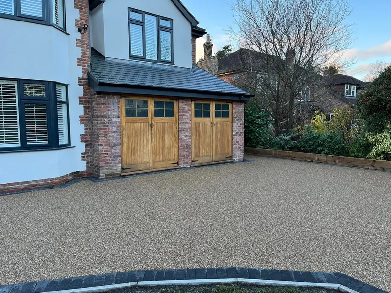 Wide sandy resin bound driveway with a charcoal sett border leading to double light wood garage doors on a red brick and white-rendered house.