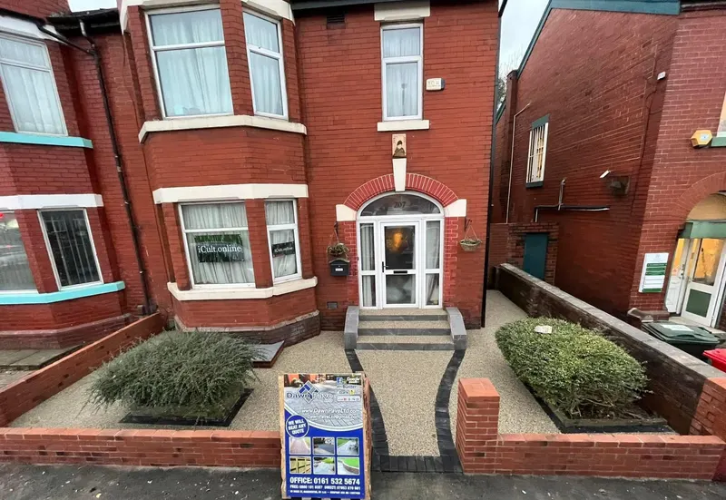Light tan resin bound front garden with dark block paving strips forming a central path and two planted beds in front of a red brick house.