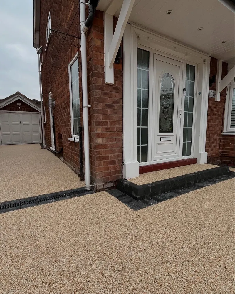 Buff resin bound entrance and side path with a block-edged front step, linear channel drain, and continuation toward a white garage.