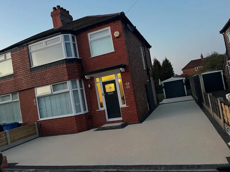 Pale cream resin bound front driveway and side run edged in dark block paving in front of a red brick house with a bay window at dusk.