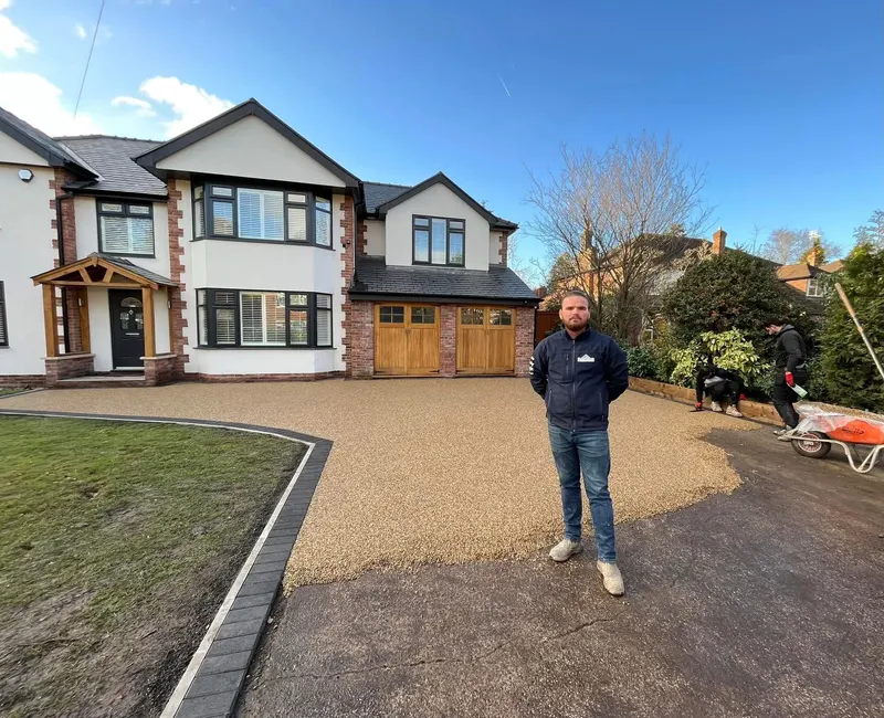Golden-buff resin bound driveway being laid with a curved charcoal block border on a white-rendered detached house while workers finish the surface.