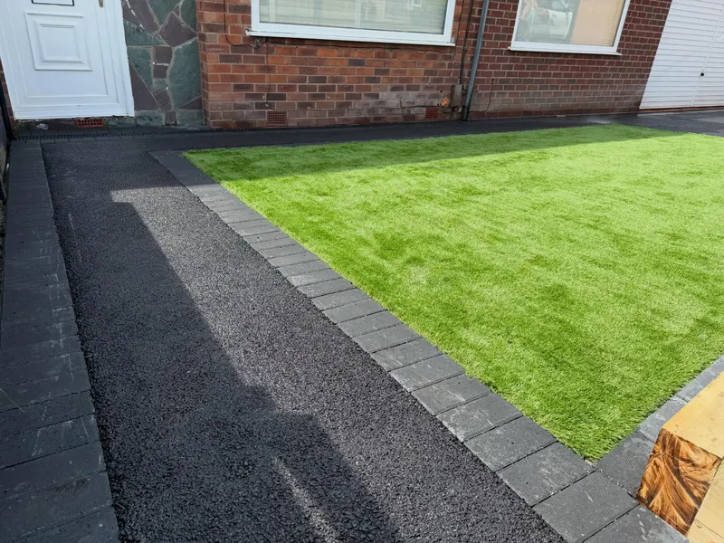 Fresh dark tarmac pathway running alongside artificial grass, both edged with charcoal block pavers against a red brick house wall.