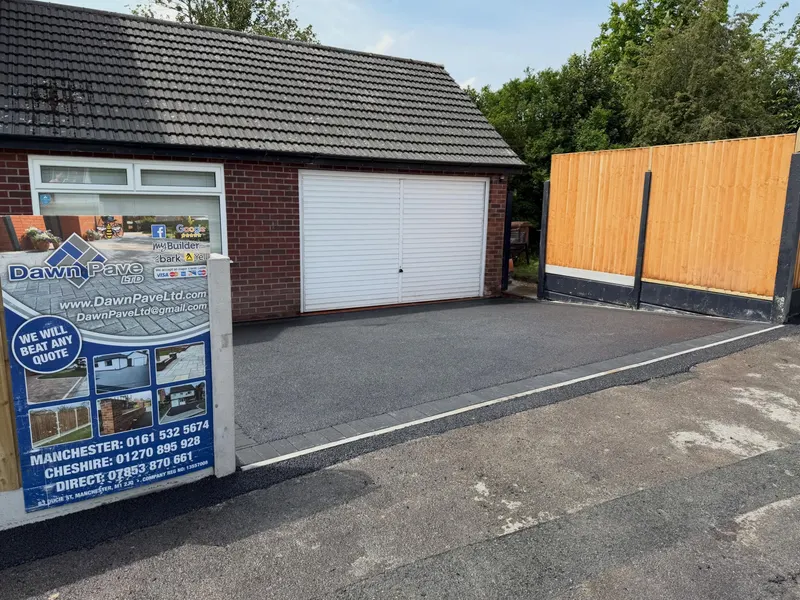 Smooth black tarmac driveway bordered by grey block paving, leading to a red brick garage with a white door and vertical-slat timber fencing.