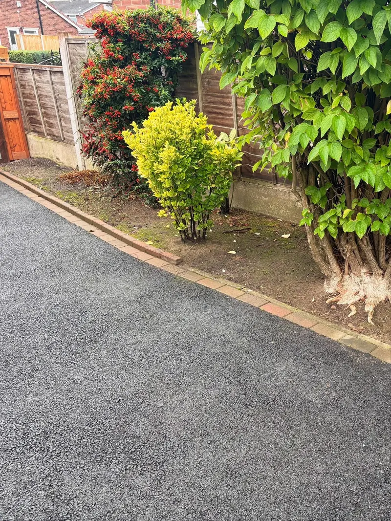 Close view of a new tarmac driveway edge lined with red and tan brick pavers, beside planted shrubs and a wooden lap-panel fence.