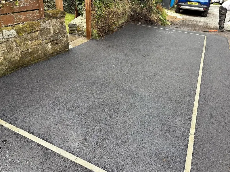 Newly laid rectangular tarmac area edged with light concrete block kerbs, next to a mossy stone wall and wooden fencing.