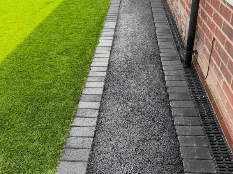 Narrow tarmac side path flanked by grey block paver edges, artificial grass on one side and a red brick wall with a linear drain on the other.