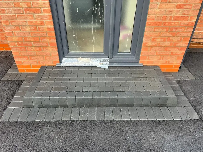 Raised charcoal block-paving doorstep and surrounding border set into smooth black tarmac at a brick wall and dark-framed glass door.