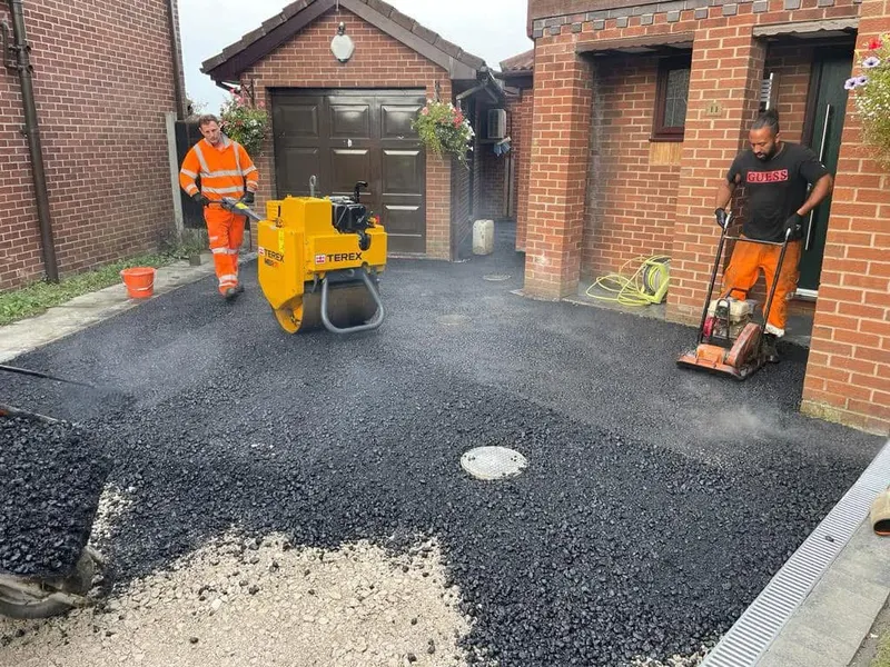 Tarmac driveway installation with workers using a walk-behind roller and plate compactor on hot black asphalt, beside a brick house and garage.