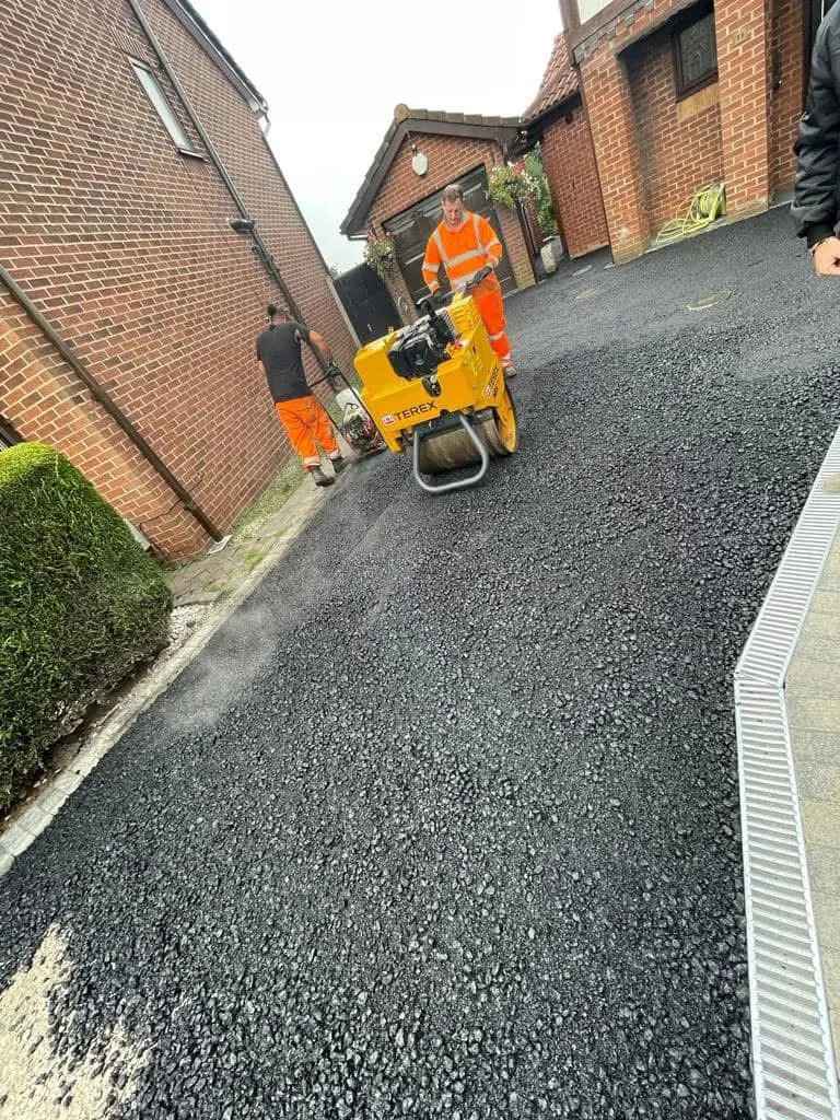 Workers in high-visibility clothing compacting steaming fresh tarmac on a residential driveway between red brick houses, with a linear drain at the edge.