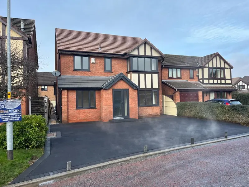 Newly laid black tarmac driveway still steaming, edged with dark block paving and bollards, in front of a mock-Tudor red brick house.