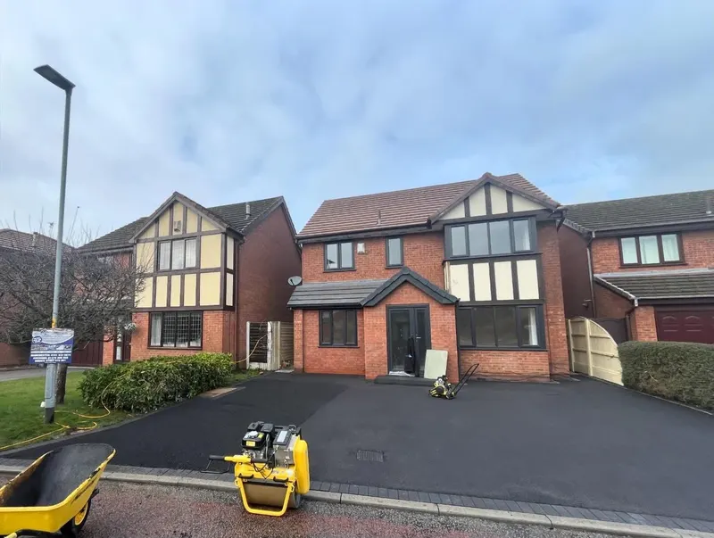 Completed black tarmac driveway with a grey block-paved front border, yellow compaction equipment still on site outside a brick and timber-frame house.