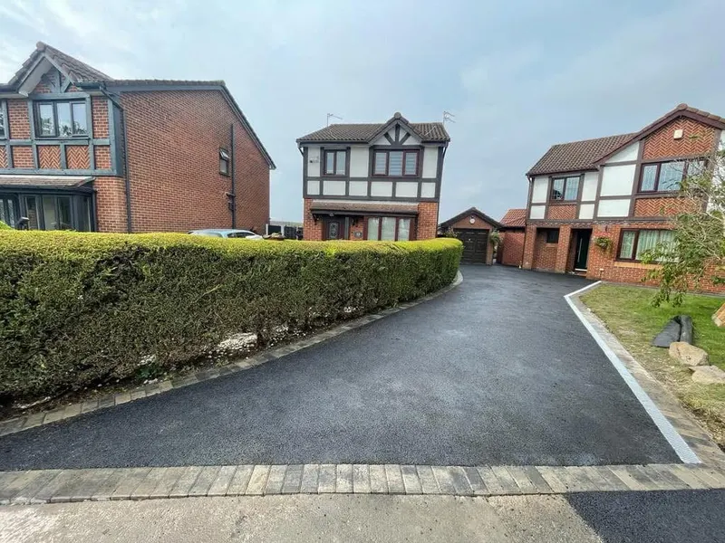 Long freshly surfaced tarmac driveway edged with light block pavers, bordered by a trimmed hedge and leading past mock-Tudor brick houses.