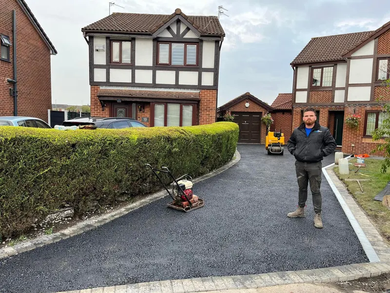 Wide black tarmac driveway being finished with a plate compactor and worker on site, edged with buff block paving beside a tall hedge.