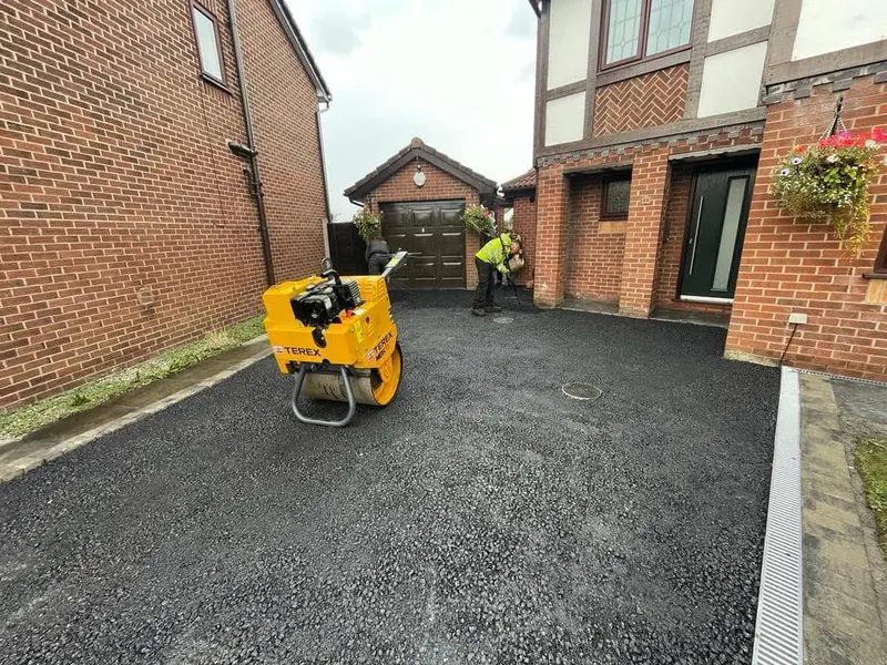 Tarmac driveway work in progress with a yellow pedestrian roller on fresh asphalt, manhole covers set in the surface, and a worker near the garage.